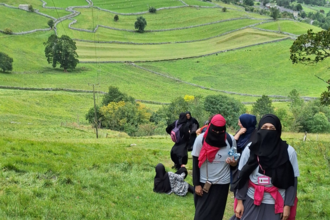 A group of hikers walking uphill on a grassy path, wearing long clothing and head coverings, with green patchwork fields, stone walls, and rolling hills stretching into the distance under a cloudy sky.
