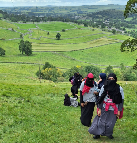 A group of hikers walking uphill on a grassy path, wearing long clothing and head coverings, with green patchwork fields, stone walls, and rolling hills stretching into the distance under a cloudy sky.